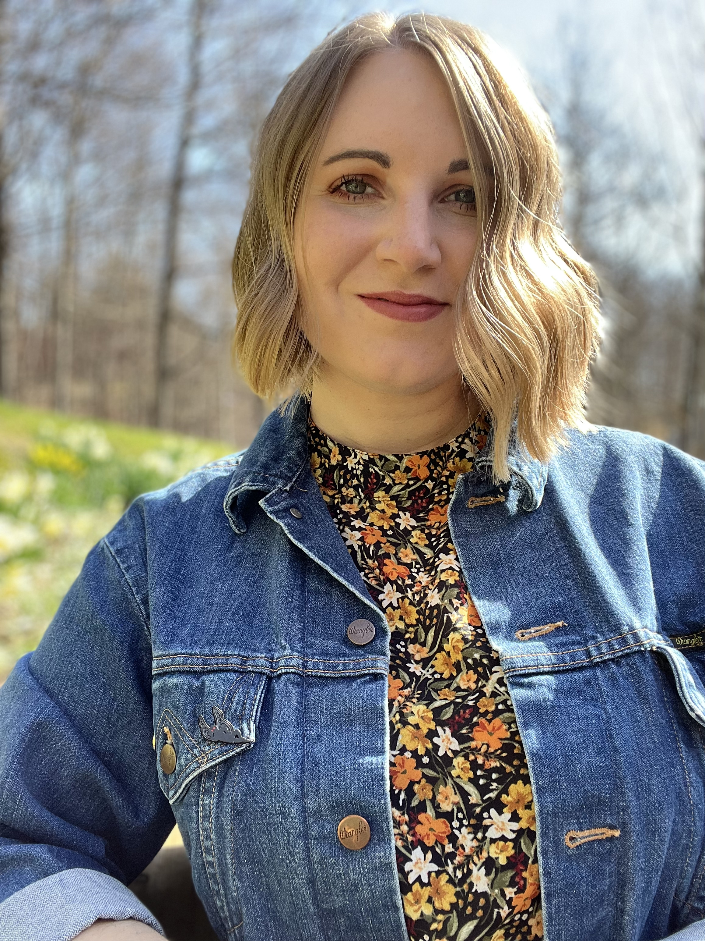 Picture of blonde woman in jean jacket and floral dress smiles with mouth closed at the camera.