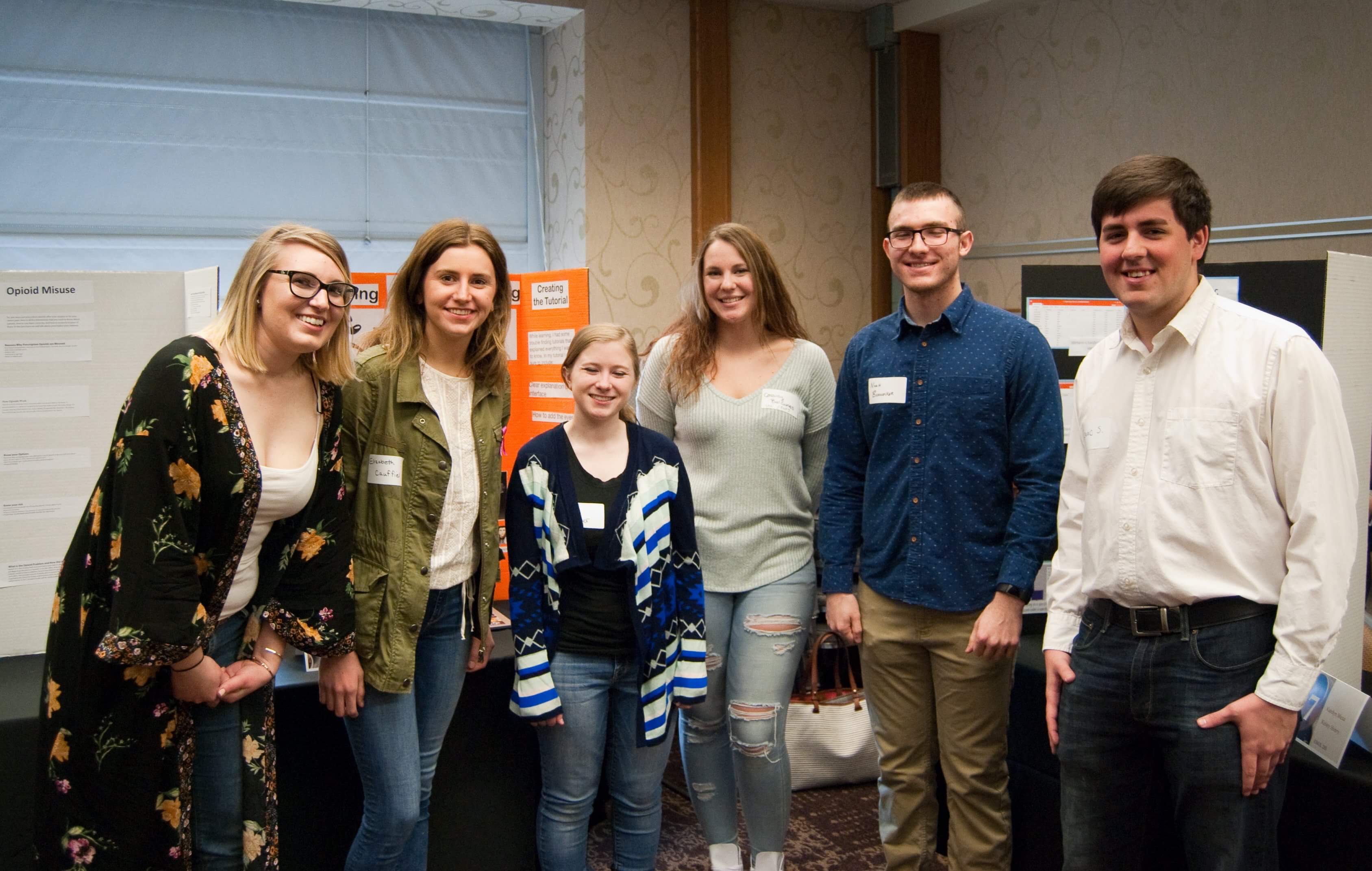 5 smiling people staring at camera in assembly hall with tri-fold poster presentations in background.