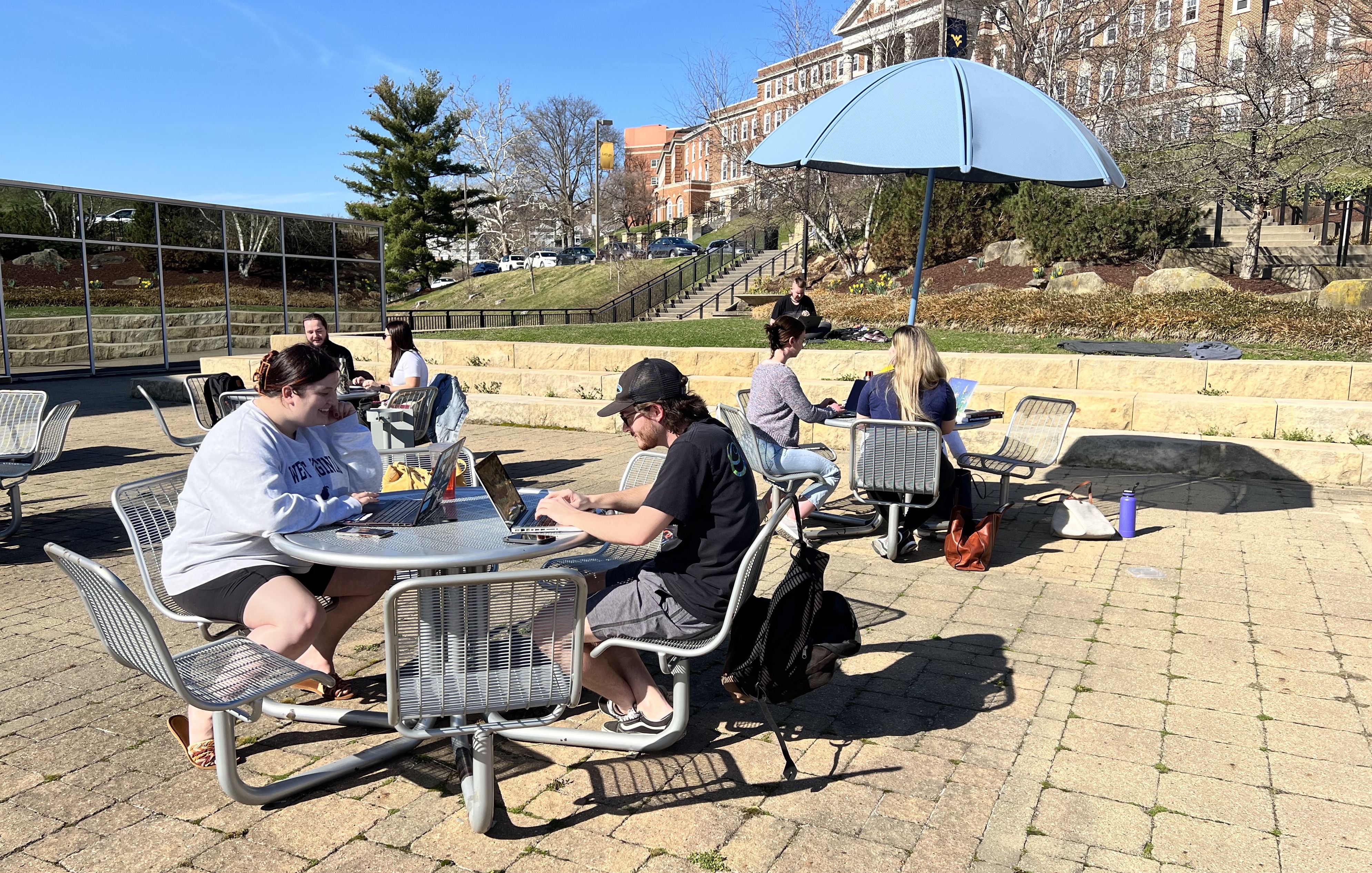 Group of students sitting outside at picnic tables on sunny spring day with computers open.